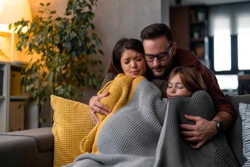 Why Isn’t My Furnace Heating My Entire Home? Photo of a family of three freezing in their home, huddled under blankets to keep warm.